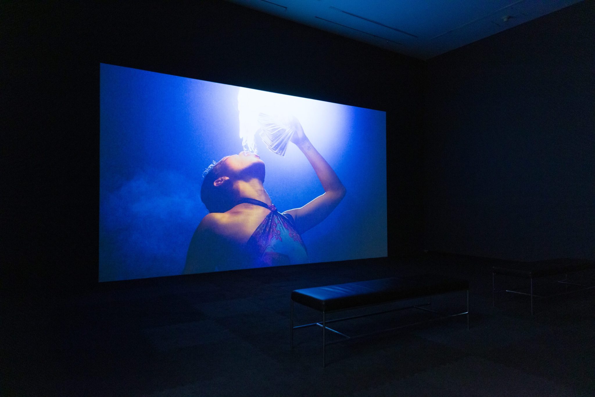 A projected image in a dark room shows a person tilting their head back and holding a bright light source to their mouth.