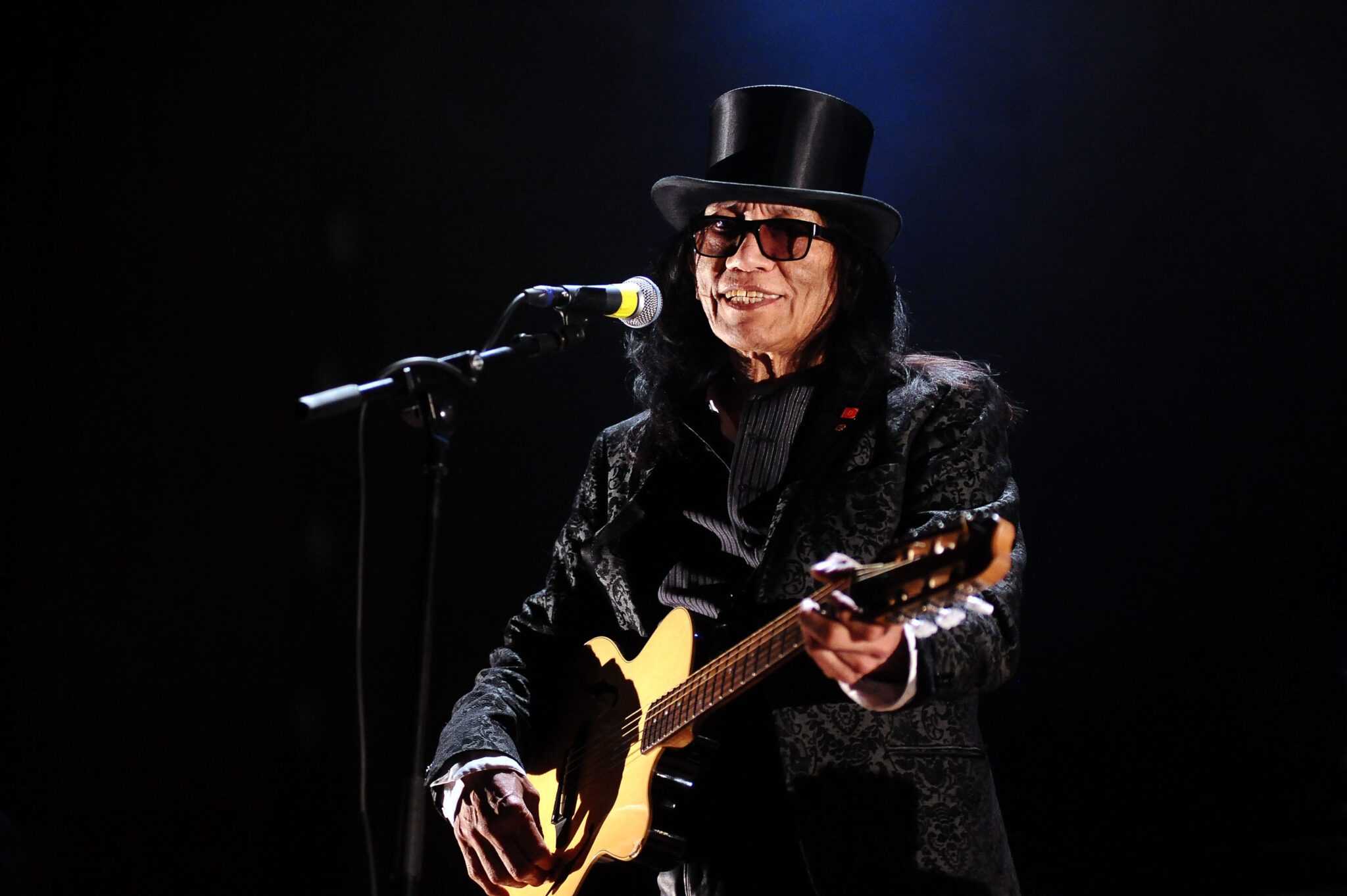 A musician in a black outfit and top hat playing an electric guitar and singing into a microphone on stage under a spotlight.