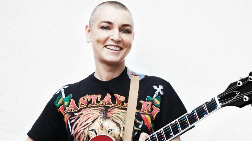 A smiling person with a shaved head, wearing a graphic t-shirt, holding a guitar against a plain backdrop.