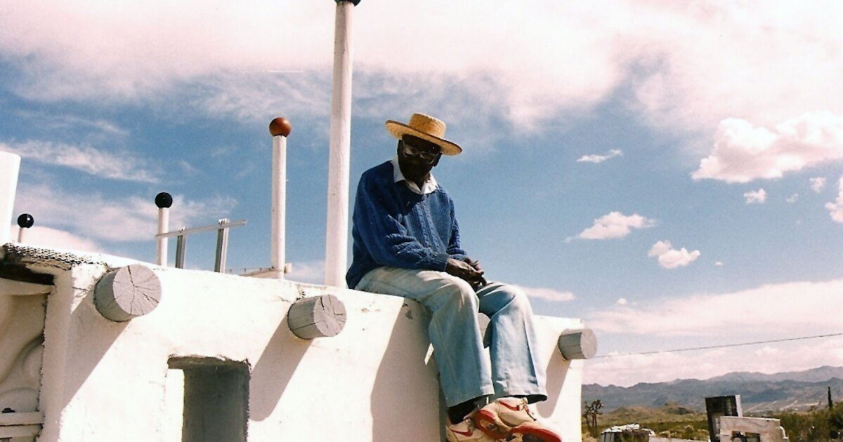 A man in a hat and sunglasses sitting pensively atop a white structure with a rural landscape in the background.