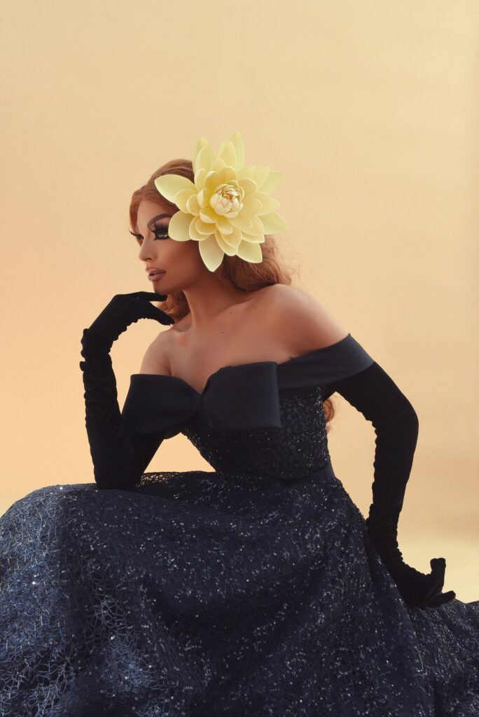 A person with dramatic makeup and a large, flower-shaped accessory adorning their hair poses elegantly against a warm-toned background.