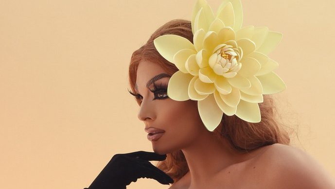 A person with dramatic makeup and a large, flower-shaped accessory adorning their hair poses elegantly against a warm-toned background.