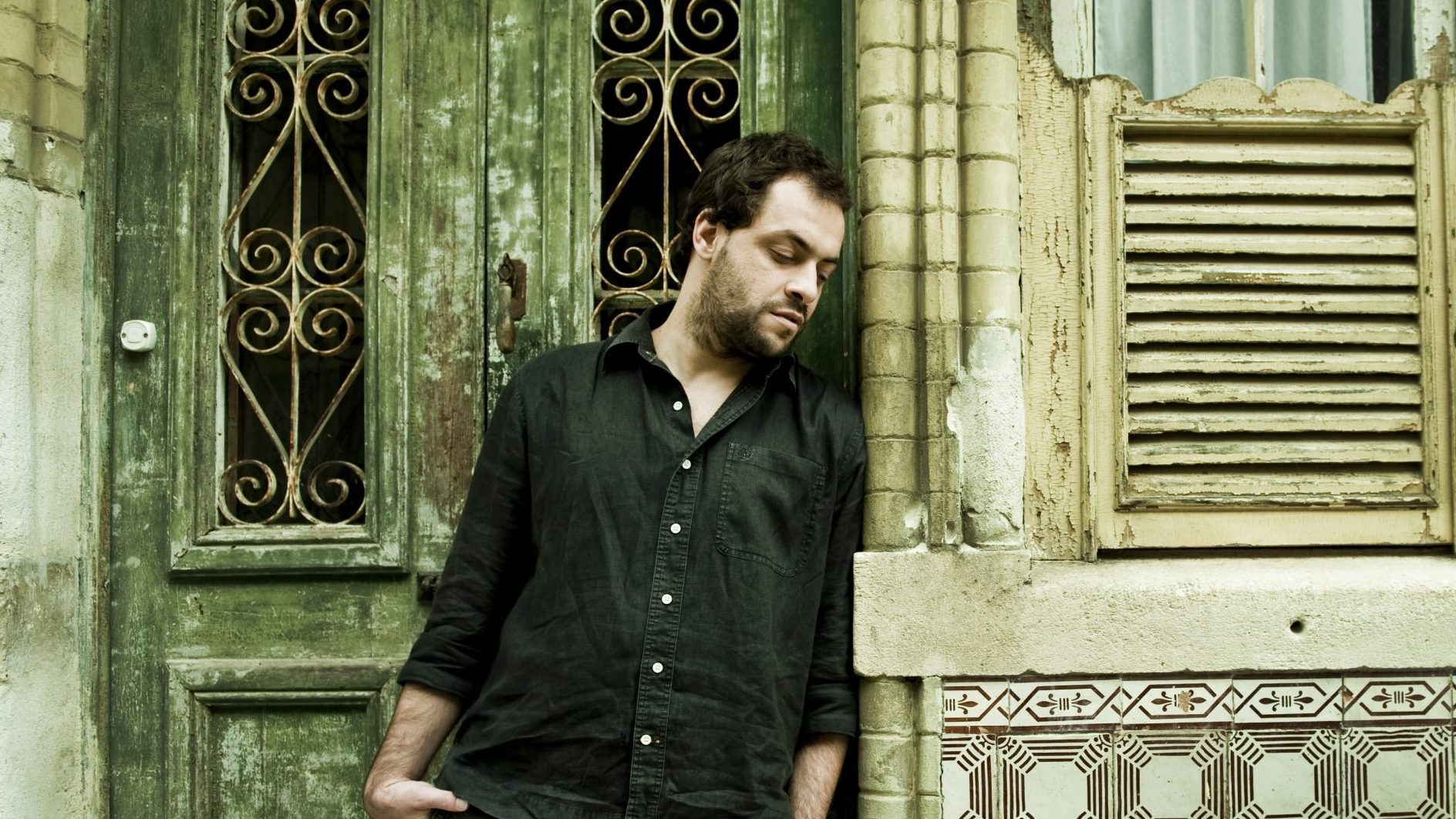 A contemplative man leaning against an old rustic wall with wrought-iron window grills and weathered shutters.