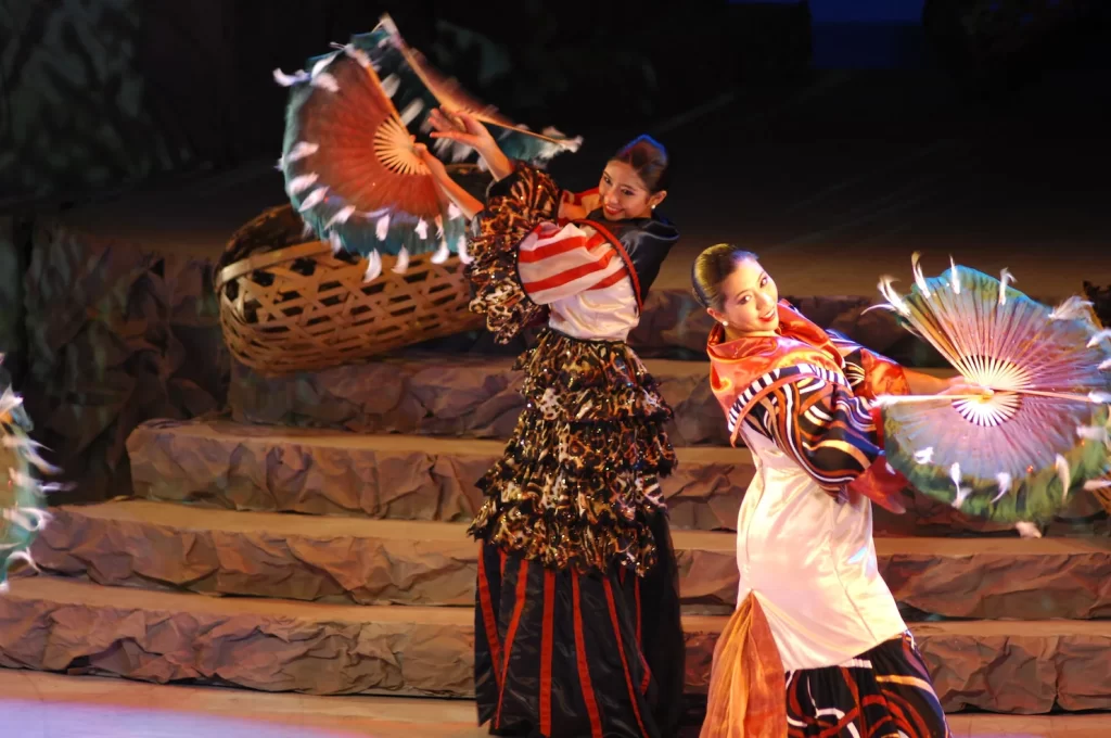 Two dancers in traditional vibrant costumes performing with large fans on a stage.