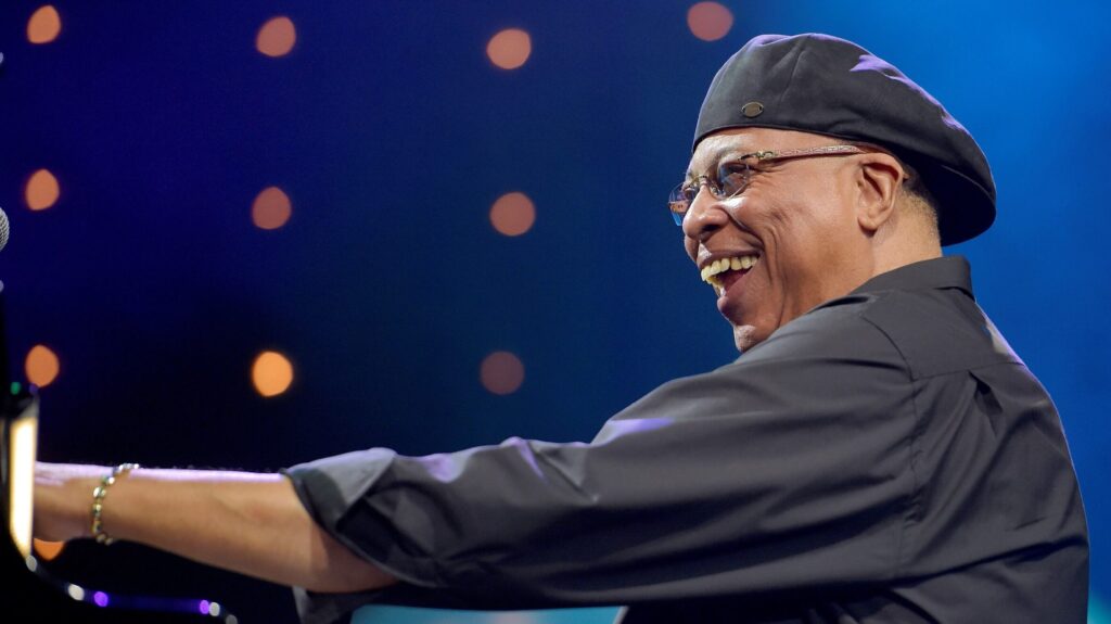 A joyful pianist wearing a beret smiles widely as he plays, surrounded by a soft glow of stage lights that create a warm, bokeh effect.
