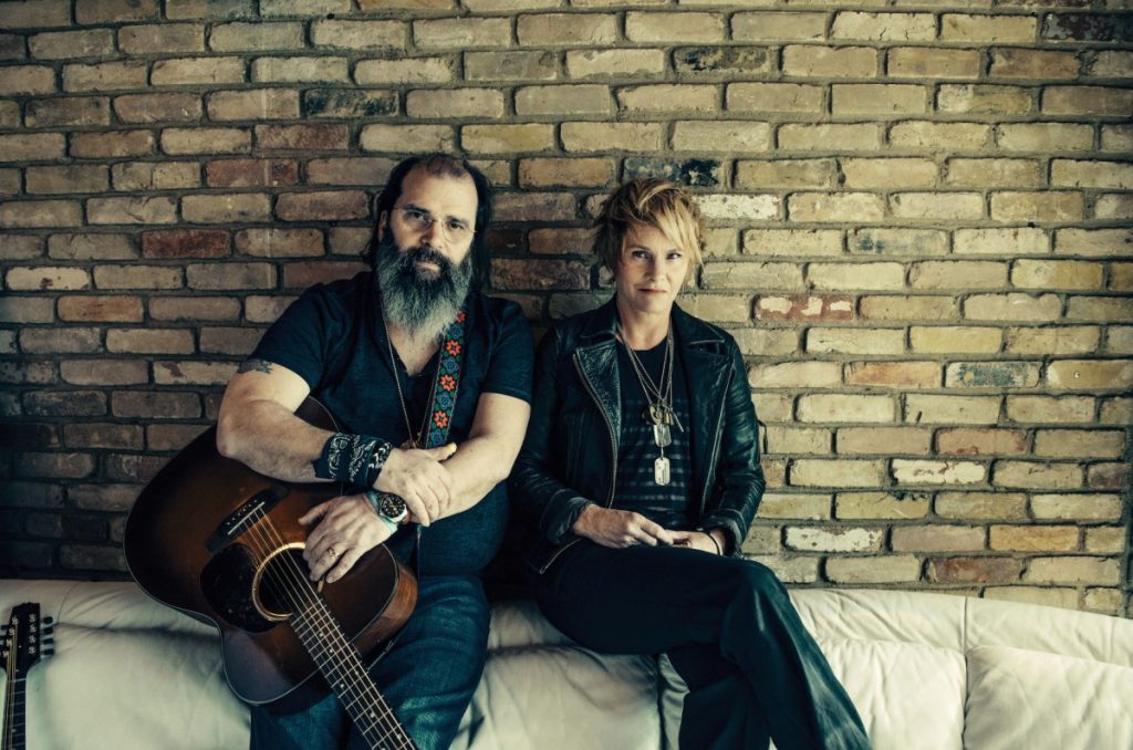 Steve Earle with a beard a guitar sitting alongside with Shawn Colvin against a rustic brick wall, both exuding a cool, artistic vibe.