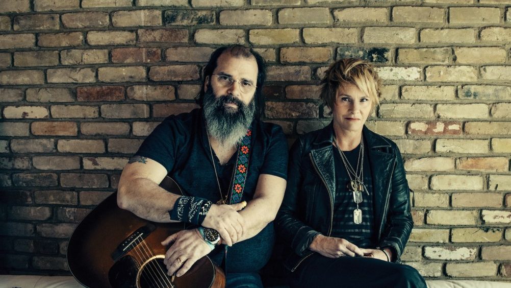 Steve Earle with a beard a guitar sitting alongside with Shawn Colvin against a rustic brick wall, both exuding a cool, artistic vibe.