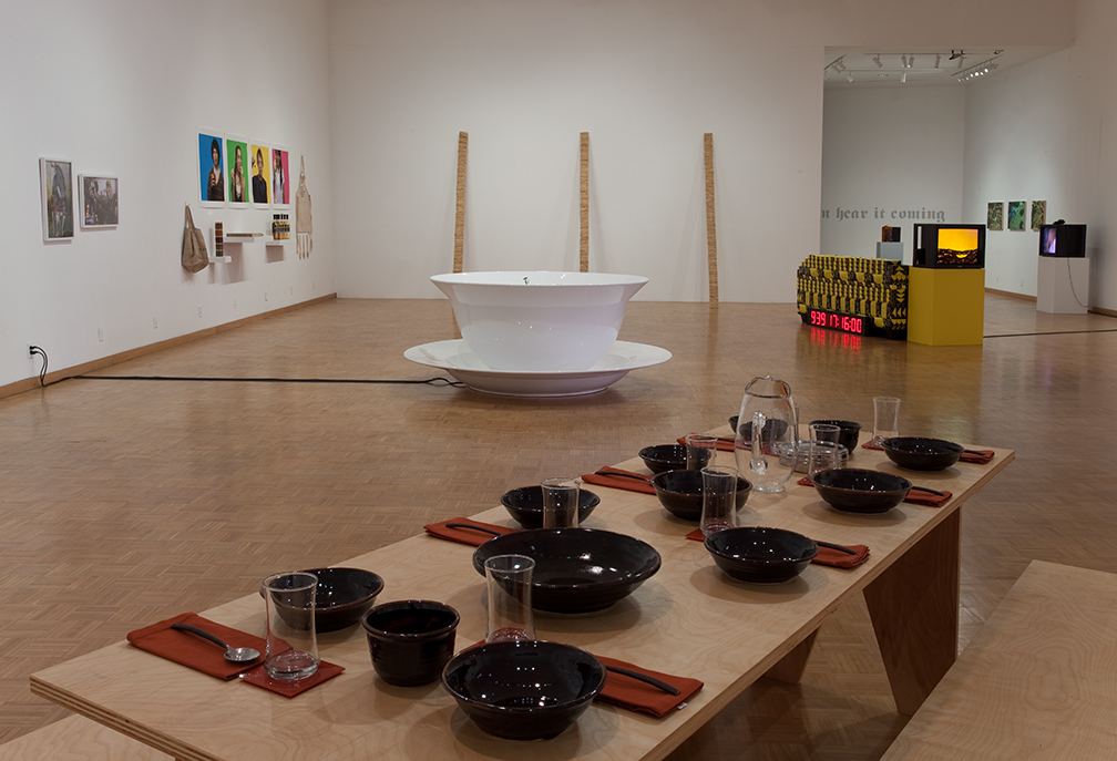 An art gallery displaying various installations, featuring a large white basin in the center surrounded by wooden poles, a table set with black bowls and glasses, and colorful wall pieces in the background.