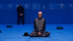 Ying Ming Tu in a dark tunic sitting cross-legged on a vibrant blue carpeted floor with a hat placed beside him, while another person stands in the background looking at framed portraits on the wall.