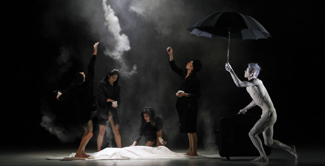 A dramatic stage performance featuring four individuals, one painted in white resembling a statue, amidst a mysterious haze with an open umbrella as a prop, conveying an intense and expressive moment.