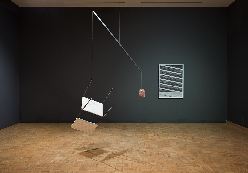 A modern art installation featuring a suspended geometric sculpture casting a shadow on the parquet flooring, juxtaposed with a framed abstract picture on a dark wall in a gallery setting.