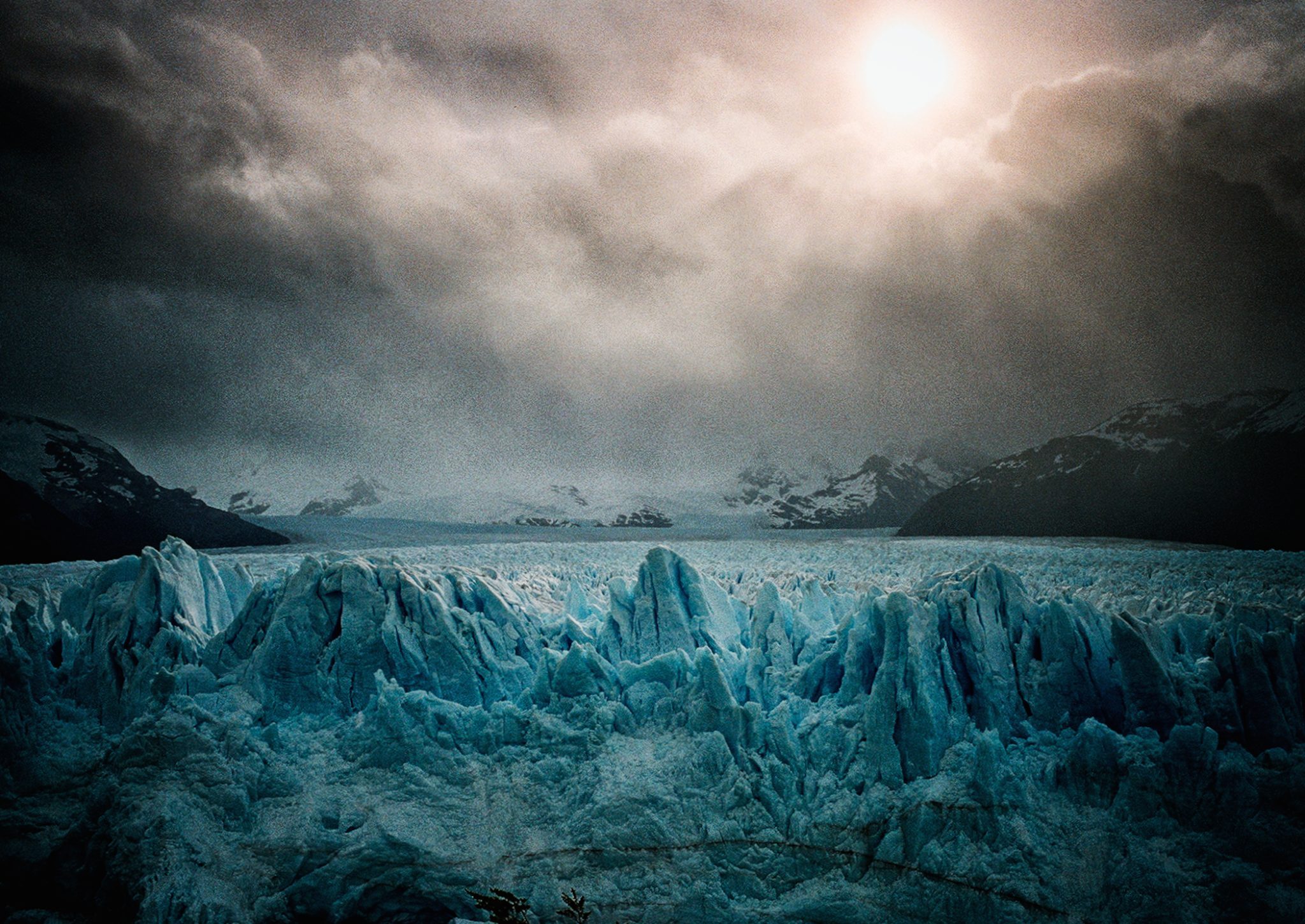 A glacier under a moody sky pierced by the sun, captured by Bruce Yonemoto.