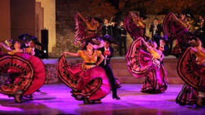 Colorful traditional mexican dance performance with dancers in vibrant swirling dresses and sombreros accompanied by a live mariachi band.