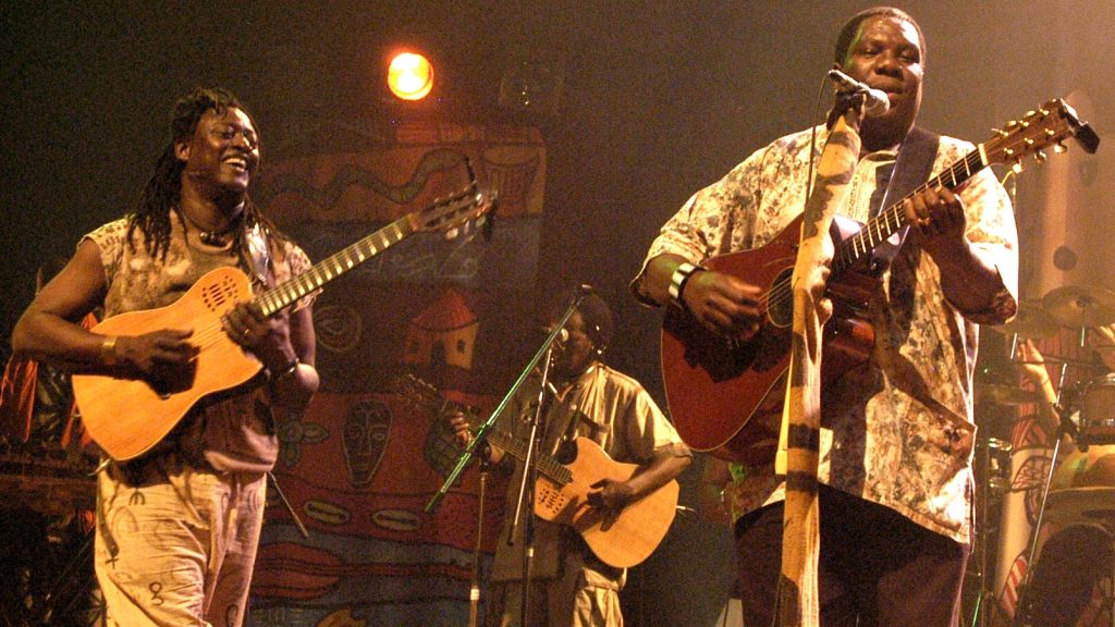 Musicians immersed in a live performance, playing acoustic guitars on stage with vibrant african-inspired backdrops and energetic stage lighting.