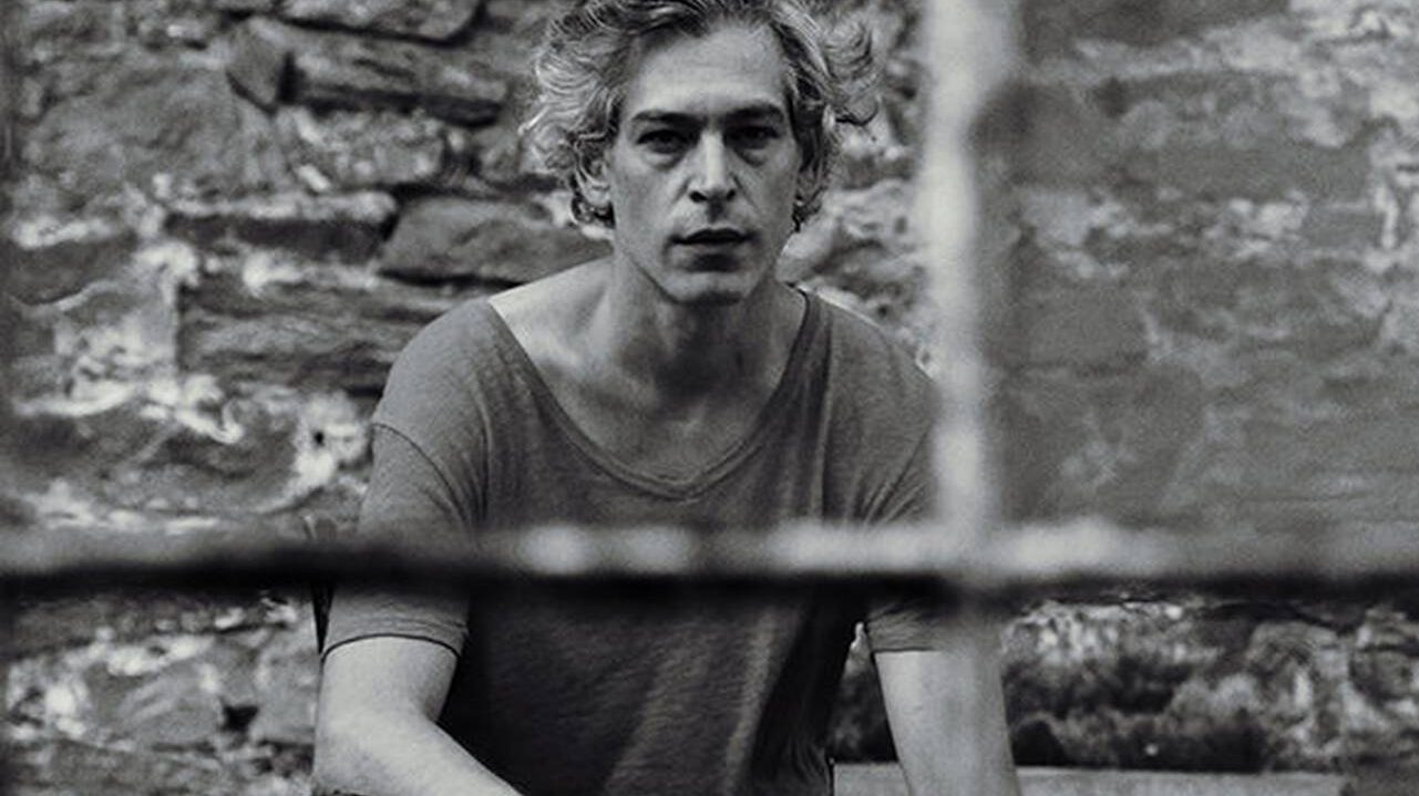 A pensively gazing man in a casual t-shirt, against a backdrop of a rustic stone wall, framed through an out-of-focus foreground.