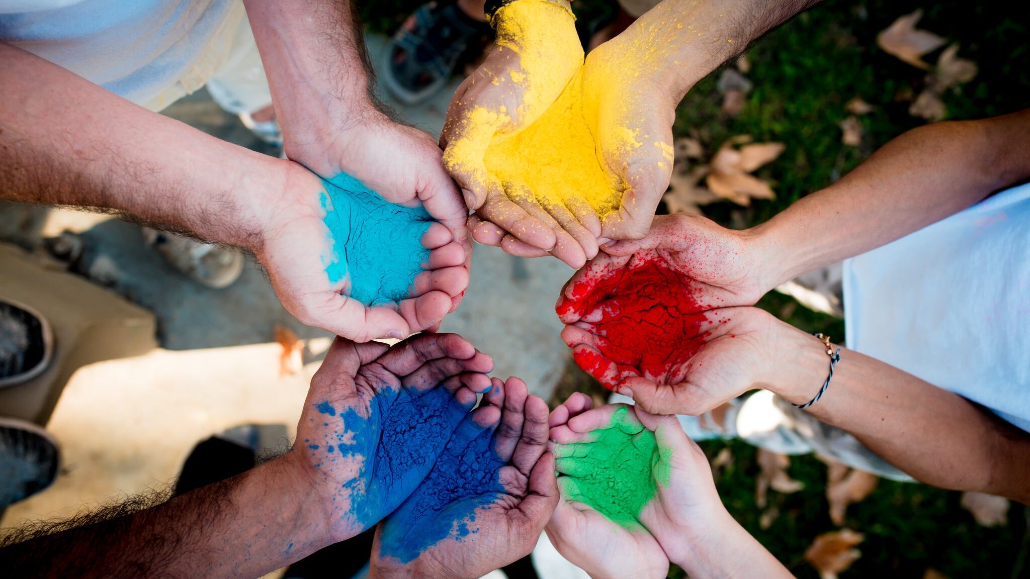 A group of people standing in a circle holding out their hands covered in vibrant colored powder, symbolizing unity, diversity, and creativity.