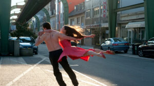 A bare-chested man in black pants lifts a woman in a flowing red dress as they perform a dramatic dance move on a sunlit urban street.