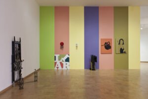 A modern art gallery with lively colored walls featuring a rainbow of vertical panels – green, yellow, blue, purple, and pink – adorned with eclectic artwork, sculptures, and some photography equipment on the wooden floor.