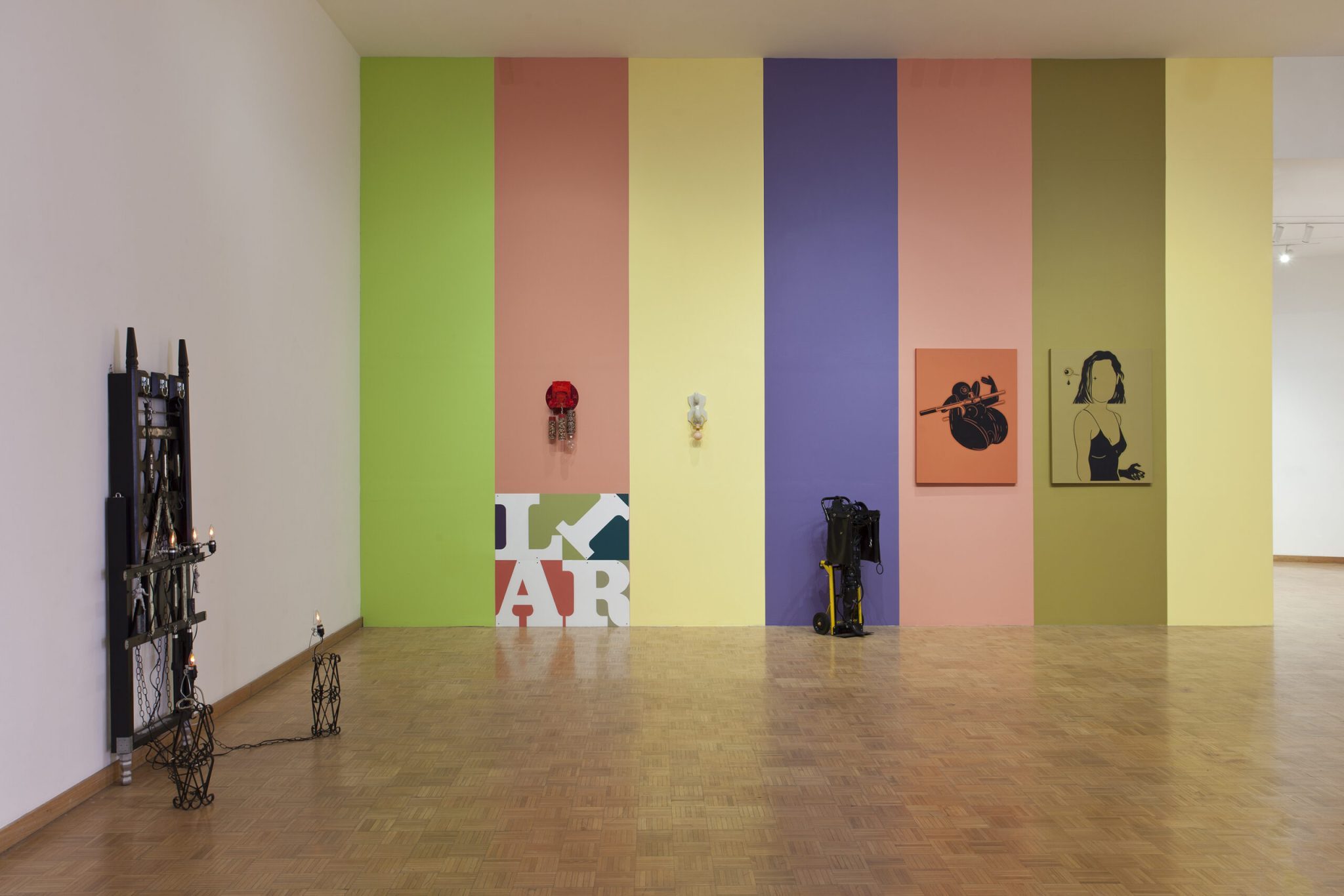 A modern art gallery with lively colored walls featuring a rainbow of vertical panels – green, yellow, blue, purple, and pink – adorned with eclectic artwork, sculptures, and some photography equipment on the wooden floor.