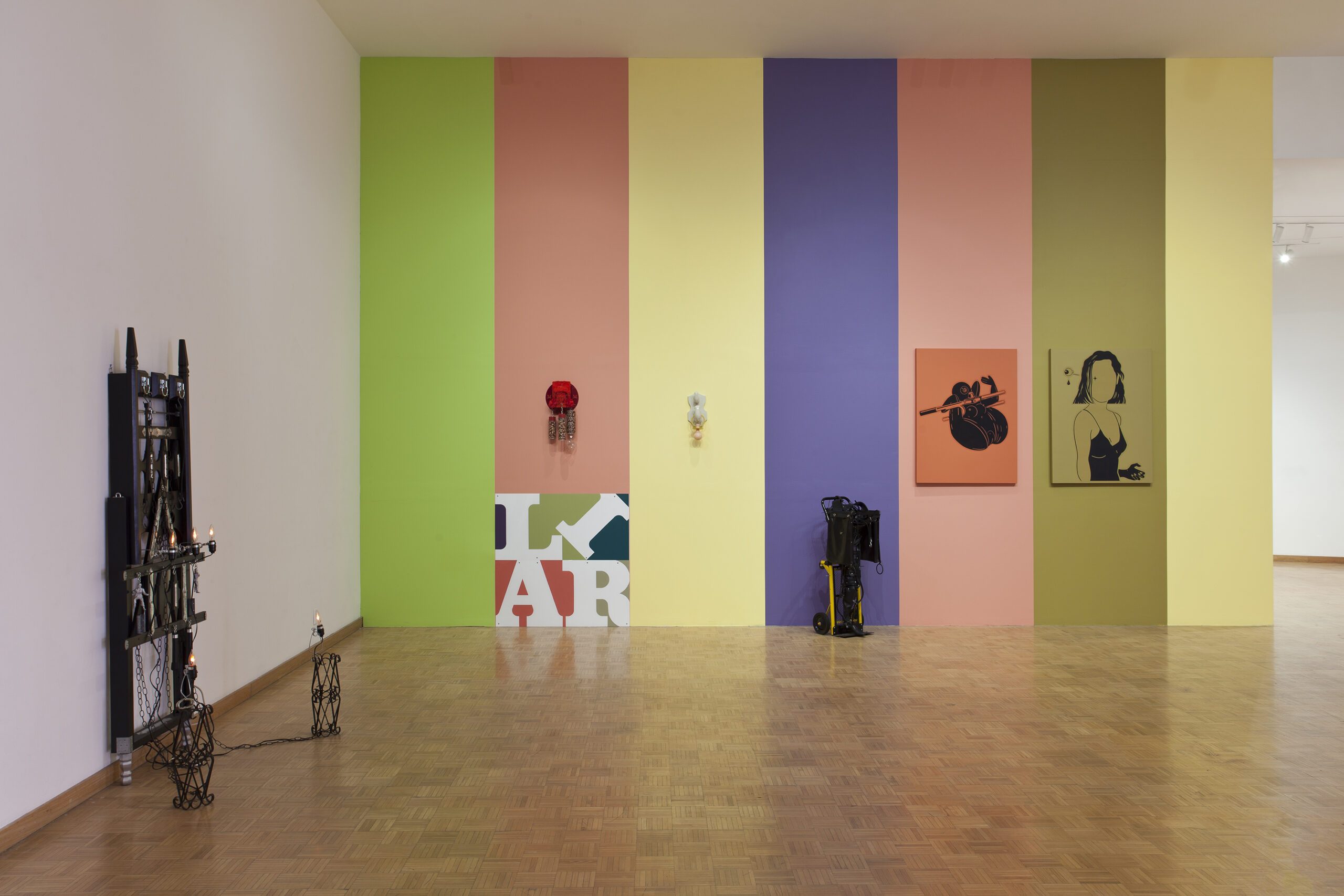 A modern art gallery with lively colored walls featuring a rainbow of vertical panels – green, yellow, blue, purple, and pink – adorned with eclectic artwork, sculptures, and some photography equipment on the wooden floor.
