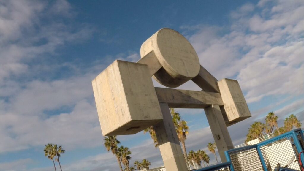 A low-angle view of a modern concrete sculpture with geometric shapes, set against a backdrop of a clear blue sky with fluffy clouds and framed by tall palm trees.
