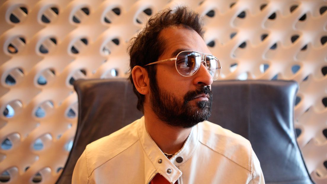 Karsh Kale wearing sunglasses and a light-colored jacket, seated against a modern geometric-patterned backdrop.