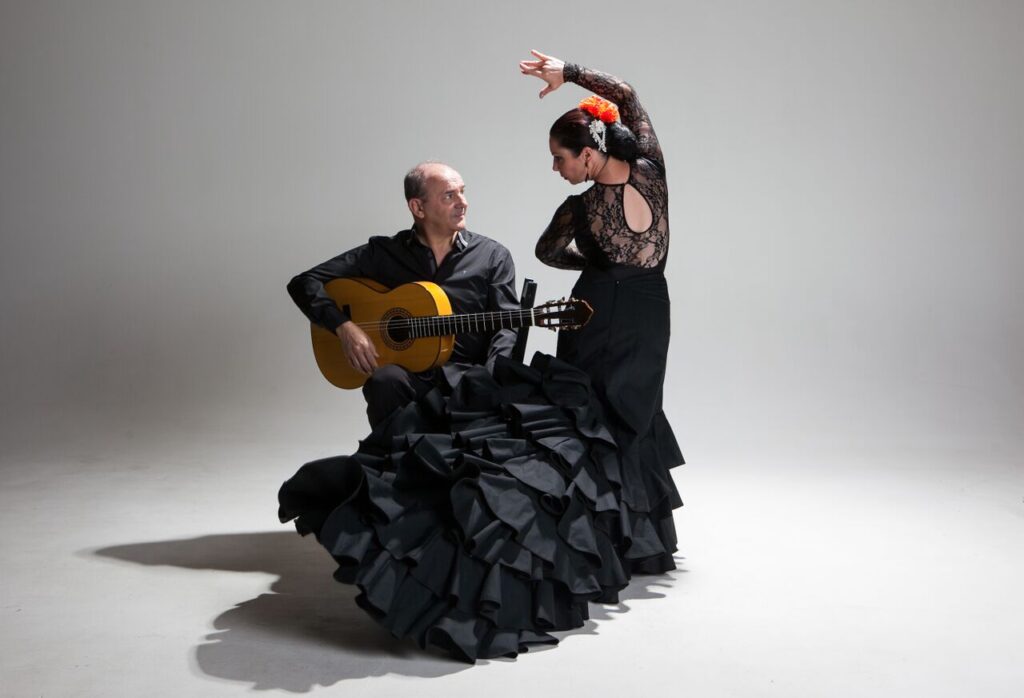 A flamenco dancer expressing emotion through her pose beside a seated guitarist who is attentively ready to play his instrument.