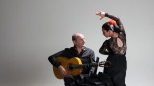 A flamenco dancer expressing emotion through her pose beside a seated guitarist who is attentively ready to play his instrument.