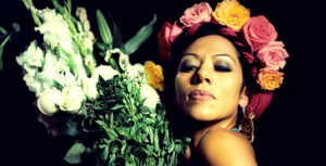 A woman with a serene expression posing with a colorful bouquet of flowers against a dark background, highlighting the vibrant beauty amidst the shadows.