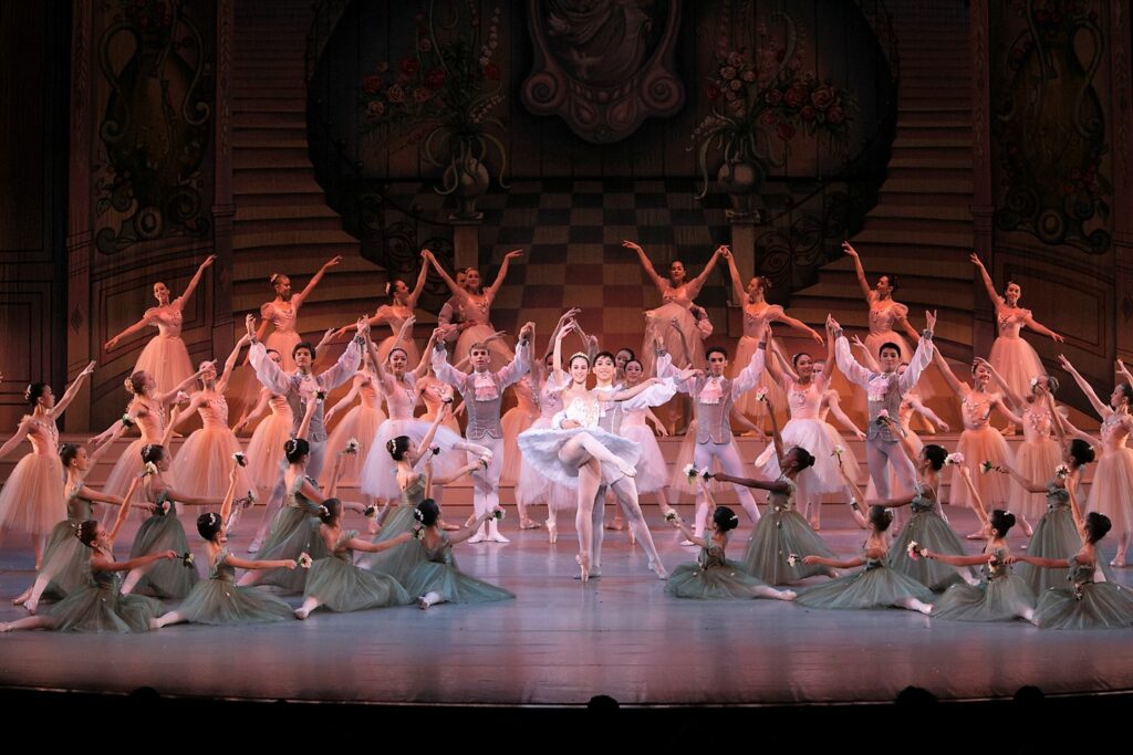 A ballet ensemble performs a graceful scene, with the principal dancers at the center in a lift, surrounded by corps de ballet in harmonious poses on a beautifully decorated stage.