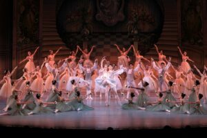 A ballet ensemble performs a graceful scene, with the principal dancers at the center in a lift, surrounded by corps de ballet in harmonious poses on a beautifully decorated stage.