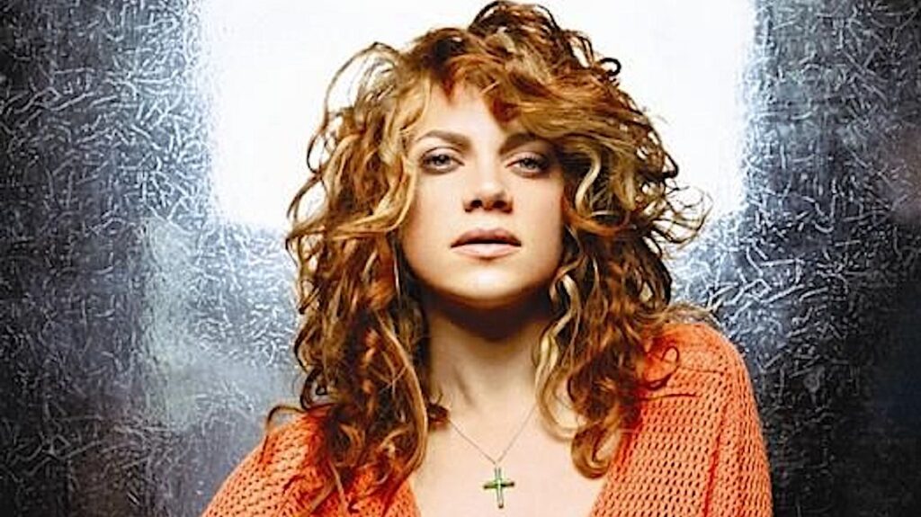 A woman with curly hair wearing an orange top and a cross necklace, against a textured backdrop.