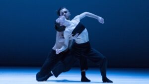 Two contemporary dancers performing an emotionally charged duet on stage, showcasing fluid movements and physical expression under blue stage lighting.