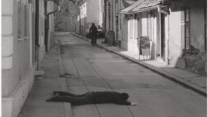 A person lying face-down on an empty street in a seemingly peaceful town, while another figure walks away in the distance.