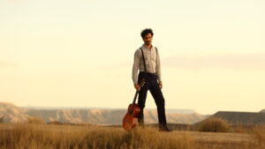 A solitary musician stands amidst a golden field, his guitar case in hand, against a backdrop of distant mesas, embodying a sense of rugged individualism and creative journey.