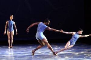 Three ballet dancers on stage in a moment of dynamic movement, with one dancer in the foreground holding another's hand, suggesting a fluid and expressive performance.