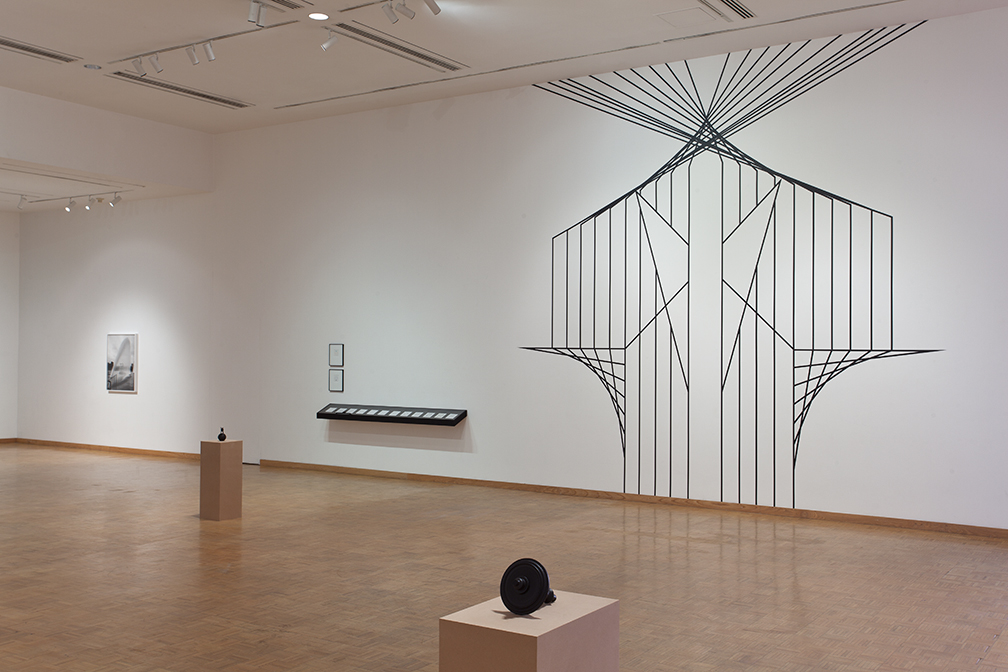 An art gallery room featuring a geometric string art installation on the wall, with other artworks and sculptures displayed on pedestals.