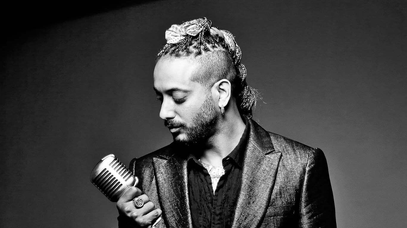Monochrome image of a stylish man holding a microphone, featuring a braided hairstyle and a unique head accessory, set against a dark background, exuding an artistic and edgy vibe.
