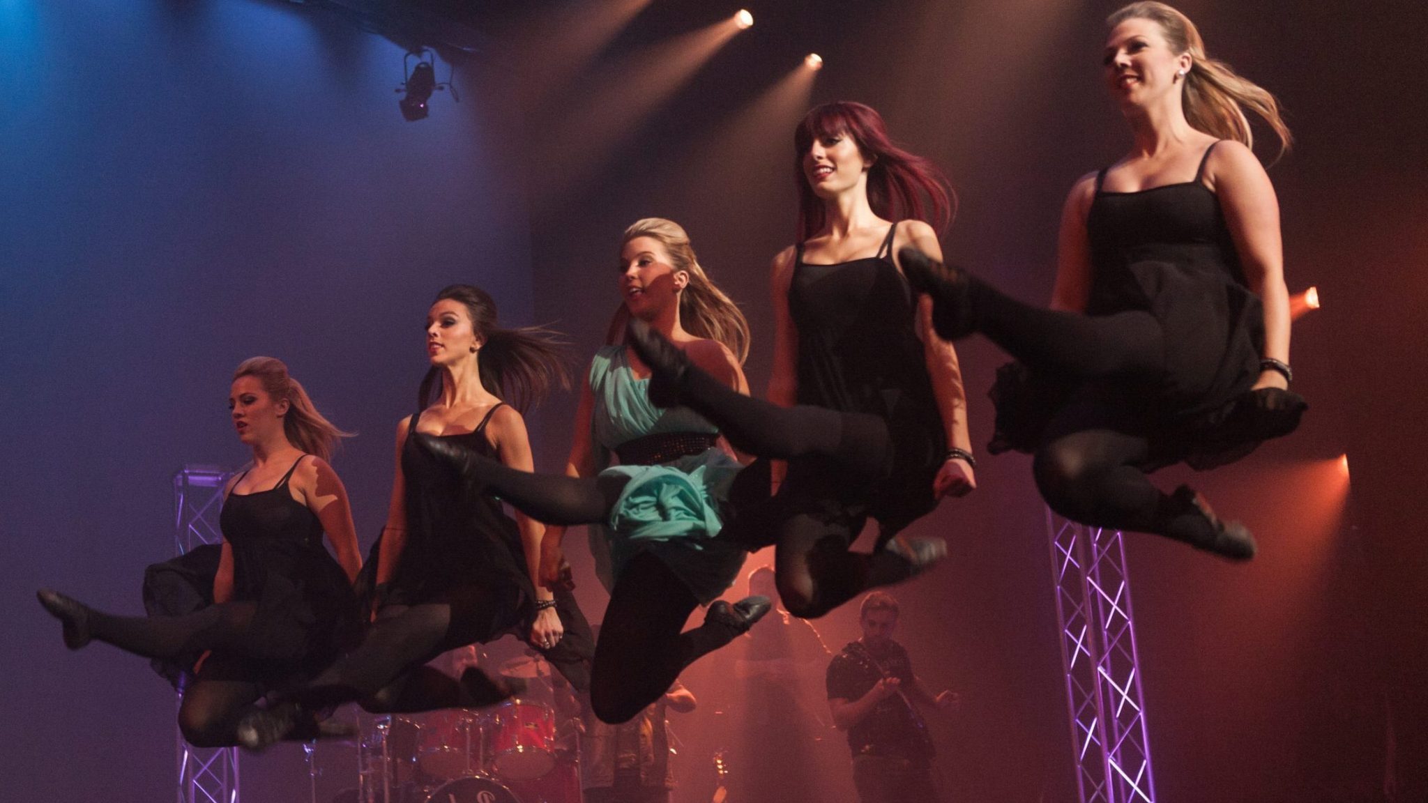 Five dancers in mid-air, showcasing a synchronized high-energy jump during a dynamic stage performance.