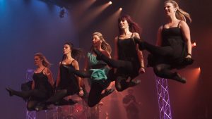 Five dancers in mid-air, showcasing a synchronized high-energy jump during a dynamic stage performance.