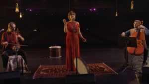 A vocalist in a red dress, Mamak Khadam, performing on stage with a cellist and a percussionist playing traditional instruments.