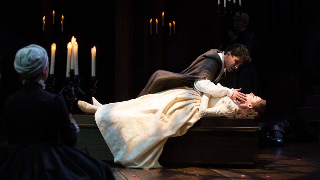 A dramatic stage scene with two actors in a period setting, where one actor tenderly holds another laying down, surrounded by candlelight, as an observer watches from the background.