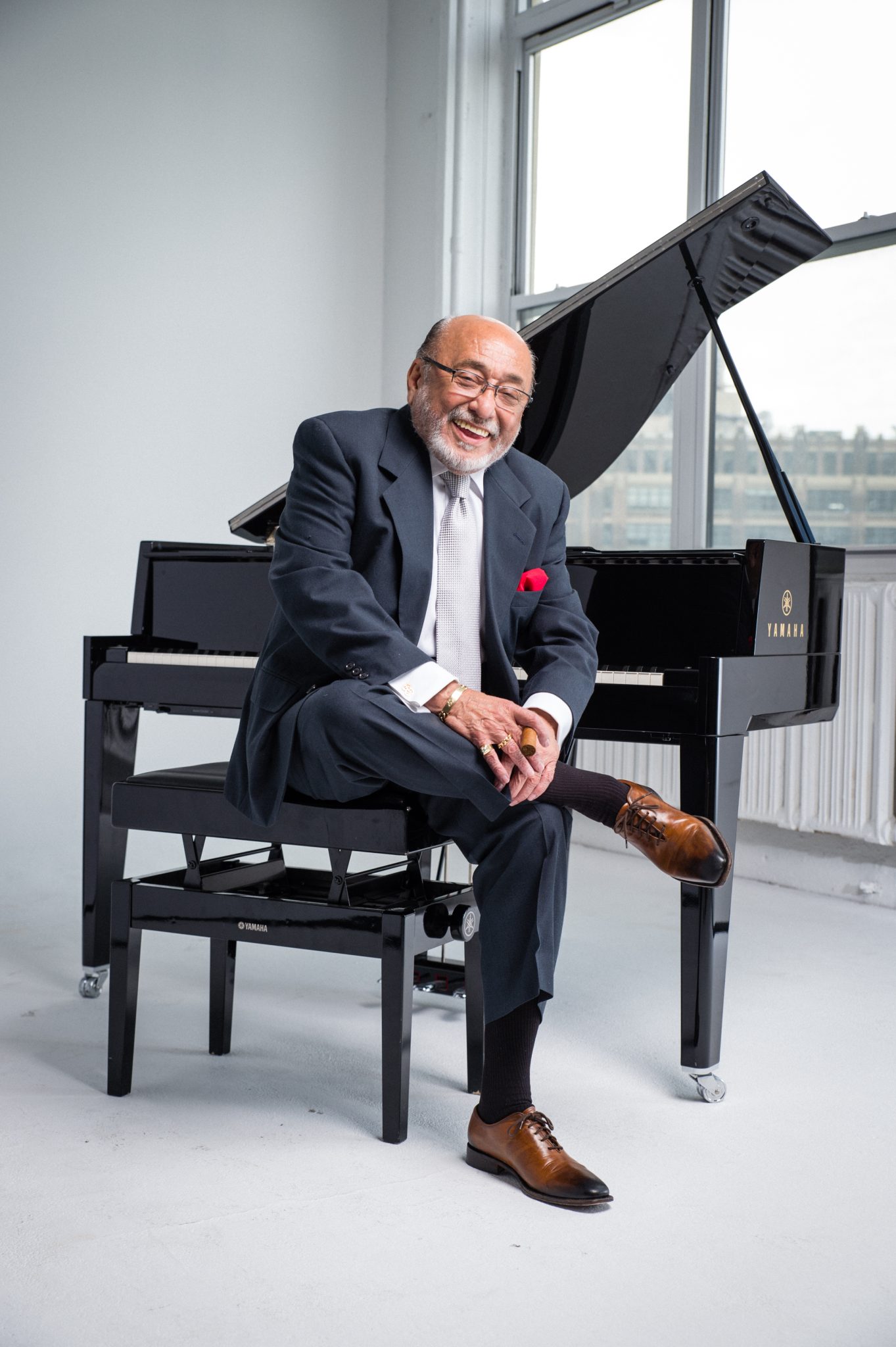 A cheerful man wearing a suit and a red pocket square sits laughing beside a grand piano in a room with a view.