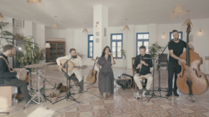 A band, Yamma Ensemble, performing in a spacious room with a vocalist, acoustic guitar, clarinet, double bass, and drum set, surrounded by a warm, bohemian interior design.