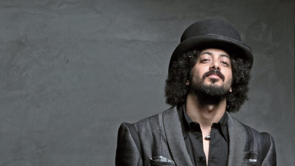 A stylish man with a distinctive afro and a bowler hat, exuding confidence against a gray backdrop.