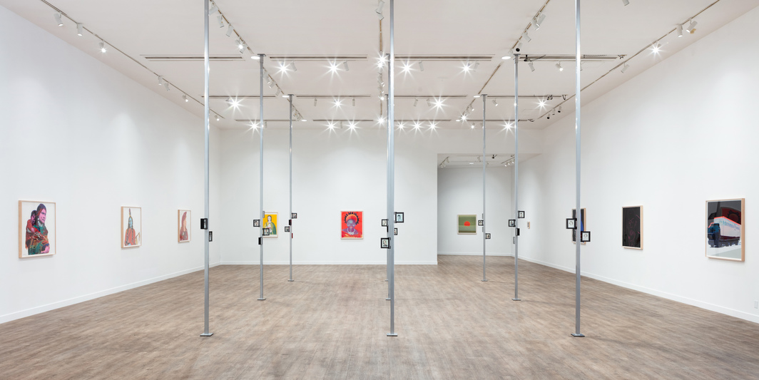 A contemporary art gallery interior displaying various paintings on white walls, with a unique installation of hanging frames in the center, under bright ceiling lights.