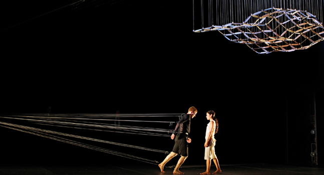 Two individuals walking alongside a dynamic light installation that creates the illusion of a floating, geometric shape through the use of strategically placed strings illuminated against a dark background.
