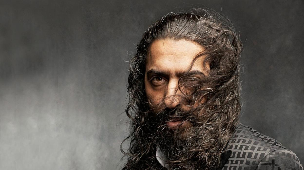 Man with long curly hair and beard against a textured gray background, exuding confidence and intensity.