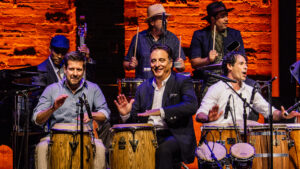 A lively band performing on stage with enthusiasm, featuring percussionists playing congas and a drum set, with a guitarist in the background, all against a vibrant brick wall backdrop.
