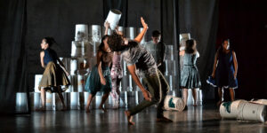 A dynamic contemporary dance performance with dancers in motion, featuring a backdrop of stacked white buckets that add an industrial aesthetic to the scene.